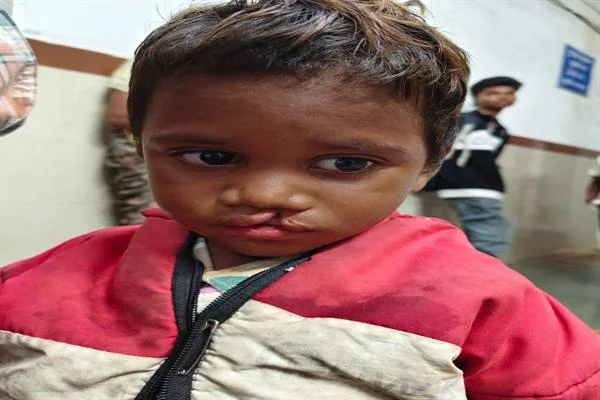 लाल और क्रीम रंग की जैकेट पहने एक छोटा बच्चा अस्पताल जैसे परिसर में खड़ा है