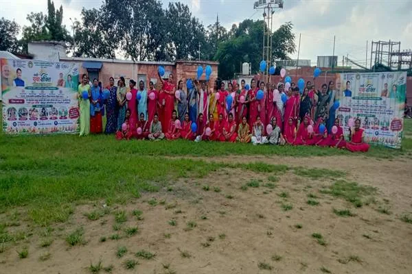 खुले मैदान में रंगीन साड़ियों में बैठी और खड़ी महिलाओं का बड़ा समूह, पीछे बैनर और नीले-सफेद गुब्बारे