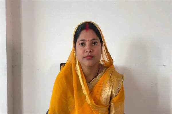 पीली साड़ी और सिर पर पल्लू डाले एक महिला सफेद दीवार के सामने बैठी हैं