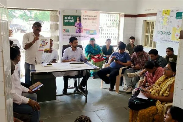 स्वास्थ्य केंद्र के कमरे में बैठे ग्रामीणों और स्वास्थ्यकर्मियों के बीच एक व्यक्ति हाथ में पुस्तिका लेकर जानकारी देता हुआ